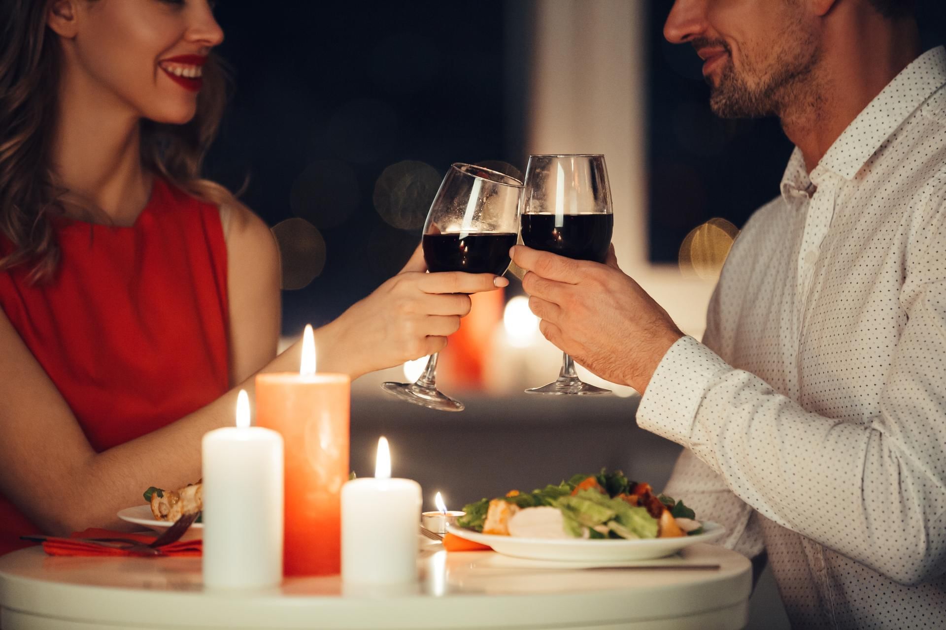 Pareja cenando y brindando con vino tinto en copas, con velas y comida en la mesa.